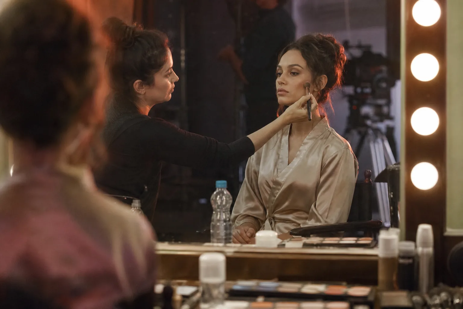 Adult woman in a satin robe in a makeup chair with bulb mirror, a makeup artist brushing her cheek before a shoot