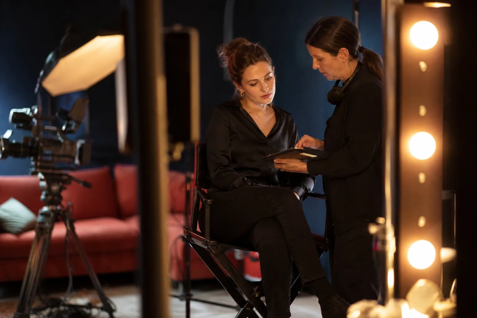 Production assistant with a clipboard talking to a dressed adult woman sitting in a makeup chair on the edge of a night film set