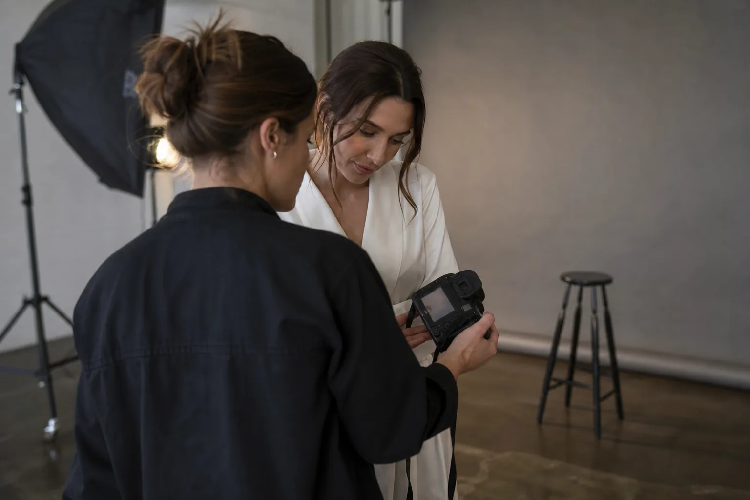 Photographer reviewing test shots on a camera with an adult woman in a robe in a photo studio with a paper backdrop