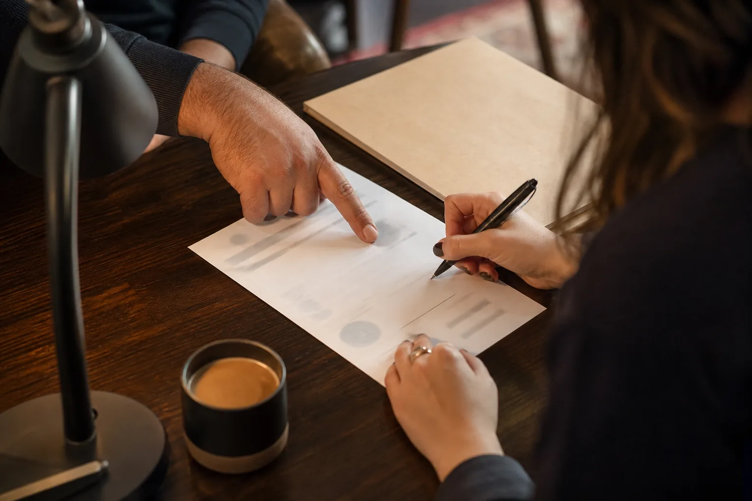 Two adults at a table, one signing a printed limits sheet with a pen while the other points to a line, in a studio office