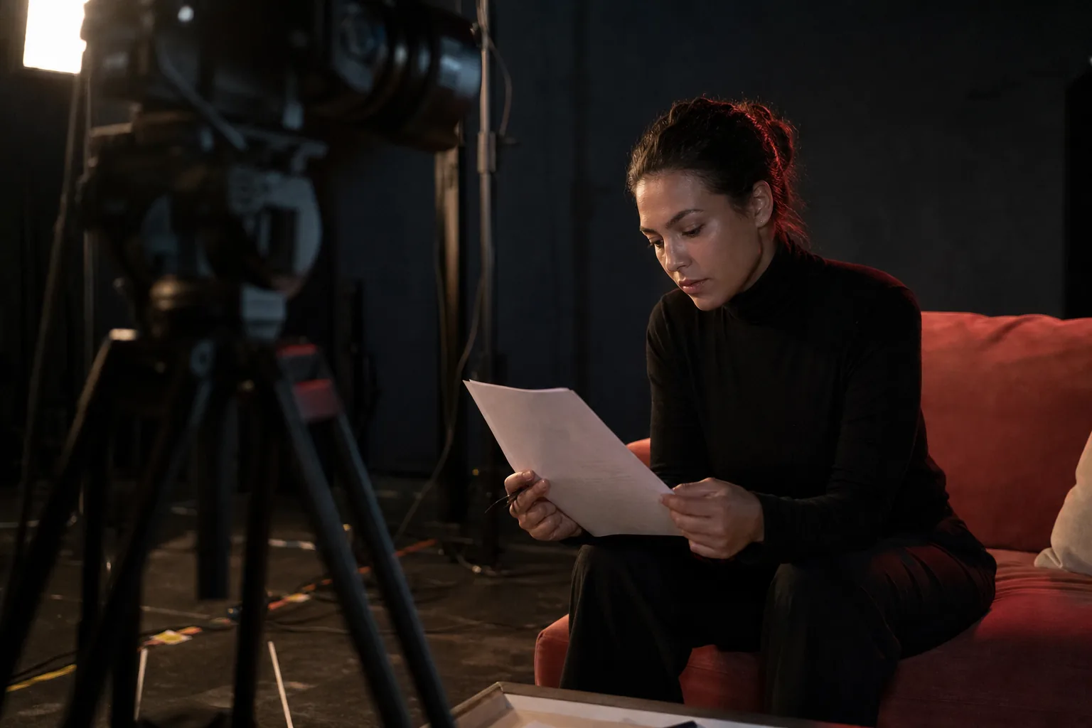 Adult woman in a black turtleneck on a coral velvet couch on a night film set, reading a one page rate agreement next to a cinema camera